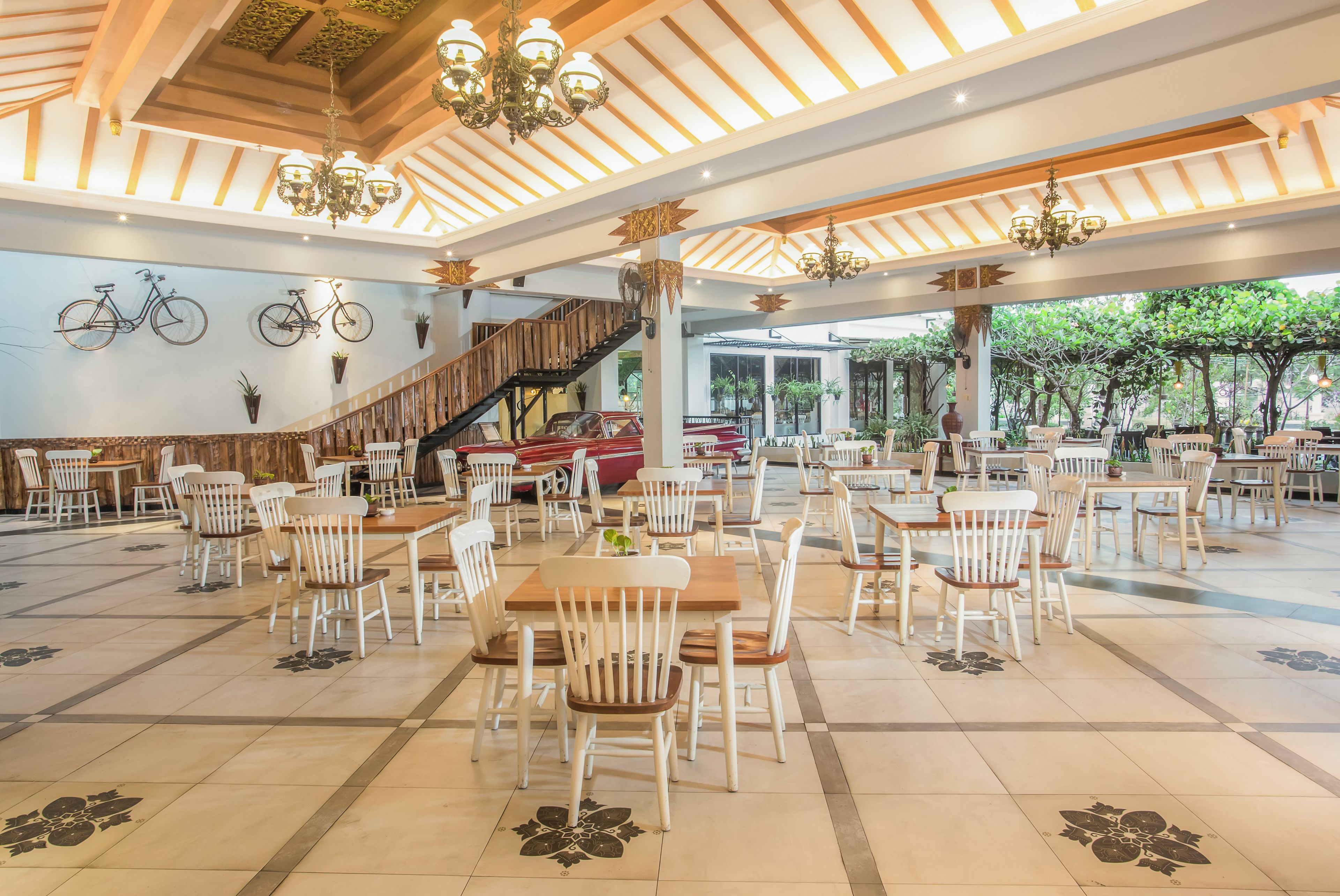 A spacious indoor dining area with wooden tables and white chairs, a staircase, and a view of greenery outside.
