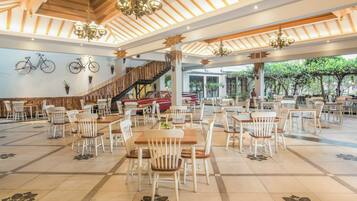 A spacious indoor dining area with wooden tables and white chairs, a staircase, and a view of greenery outside.