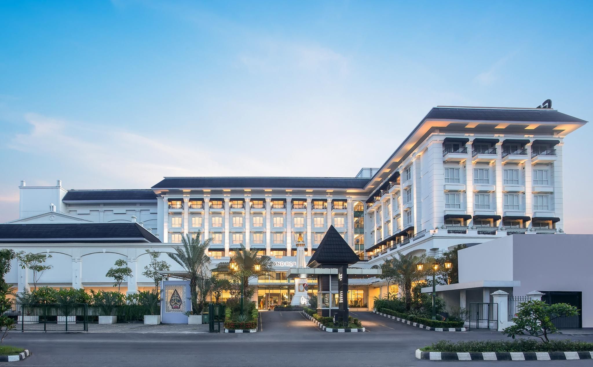 A large, multi-story hotel with a grand entrance, featuring a well-maintained garden and a prominent entrance gate.