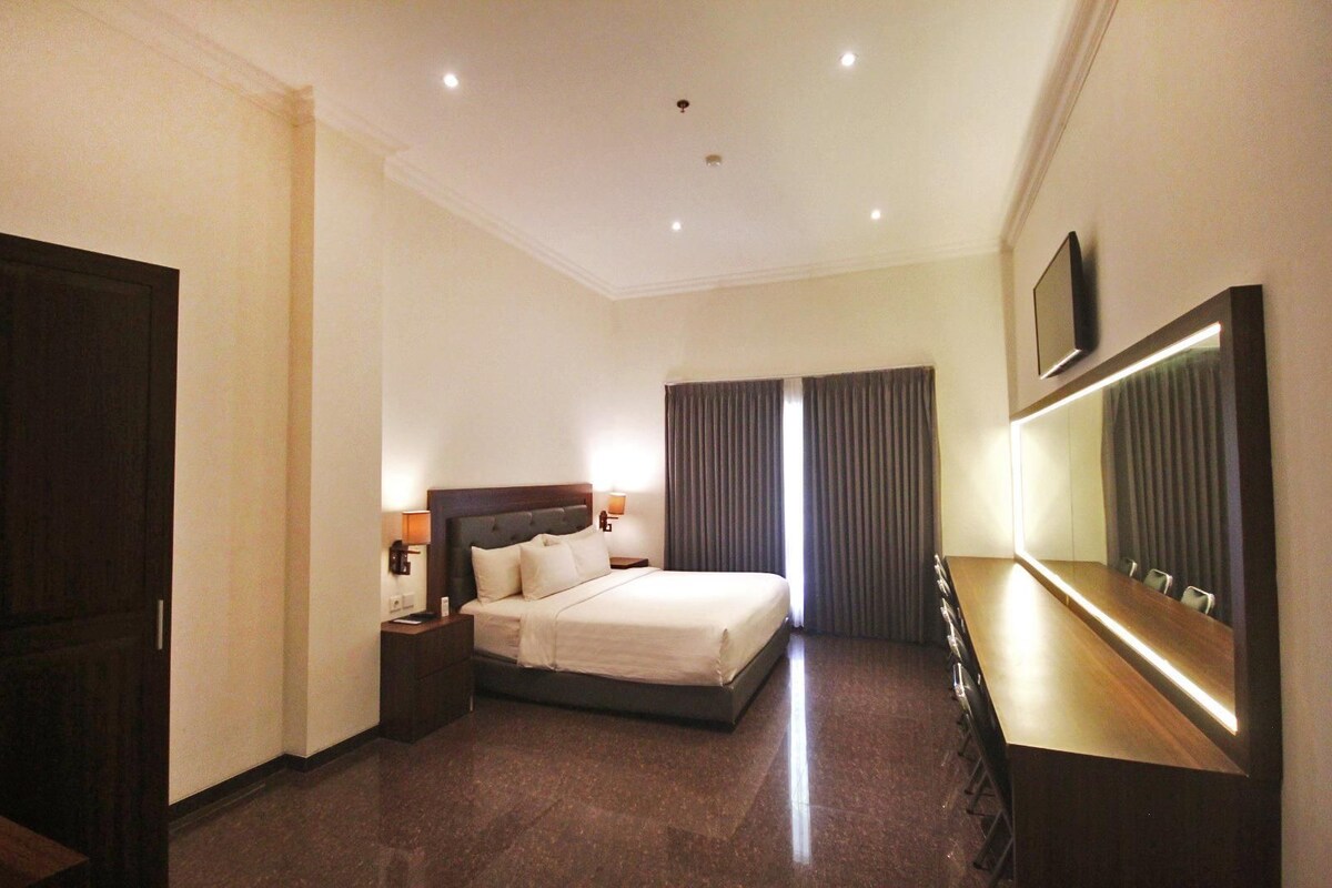 A hotel room with a bed, bedside tables, a desk with chairs, a large mirror, and a TV on the wall.