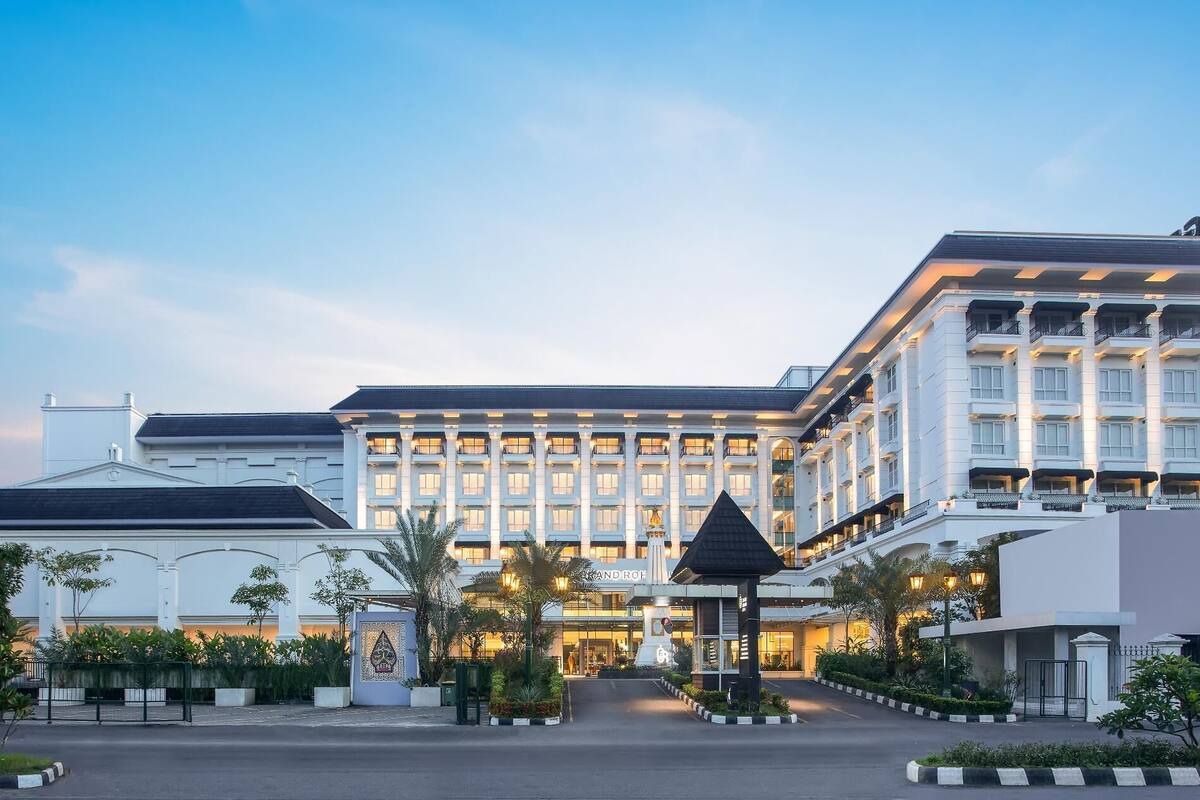 Um hotel grande, com vários andares e uma entrada imponente, com um jardim bem cuidado e um portão de entrada destacado.
