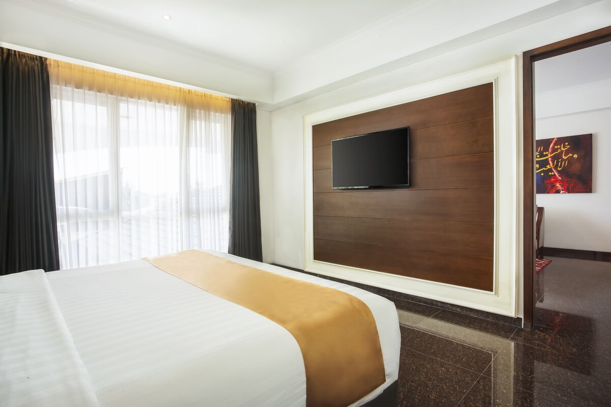 A hotel room with a large bed, a flat-screen TV mounted on a wooden panel, and a window with sheer curtains.