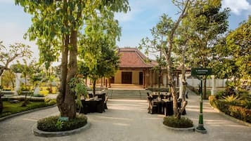 A traditional building with a tiled roof, surrounded by trees and a paved area with tables and chairs.