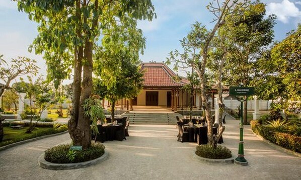 A traditional building with a tiled roof, surrounded by trees and a paved area with tables and chairs. - Grand Rohan Jogja (Banguntapan)