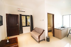 A modern living room with a sofa, a small table with a vase, a kitchen area with a sink, and a view through a window.