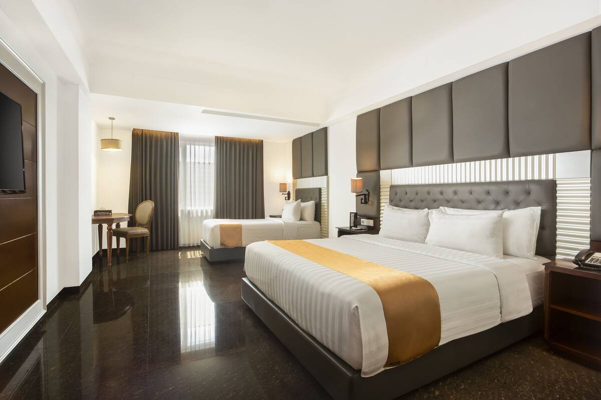 Quarto de hotel com uma cama grande, uma cama menor, uma escrivaninha e uma televisão.