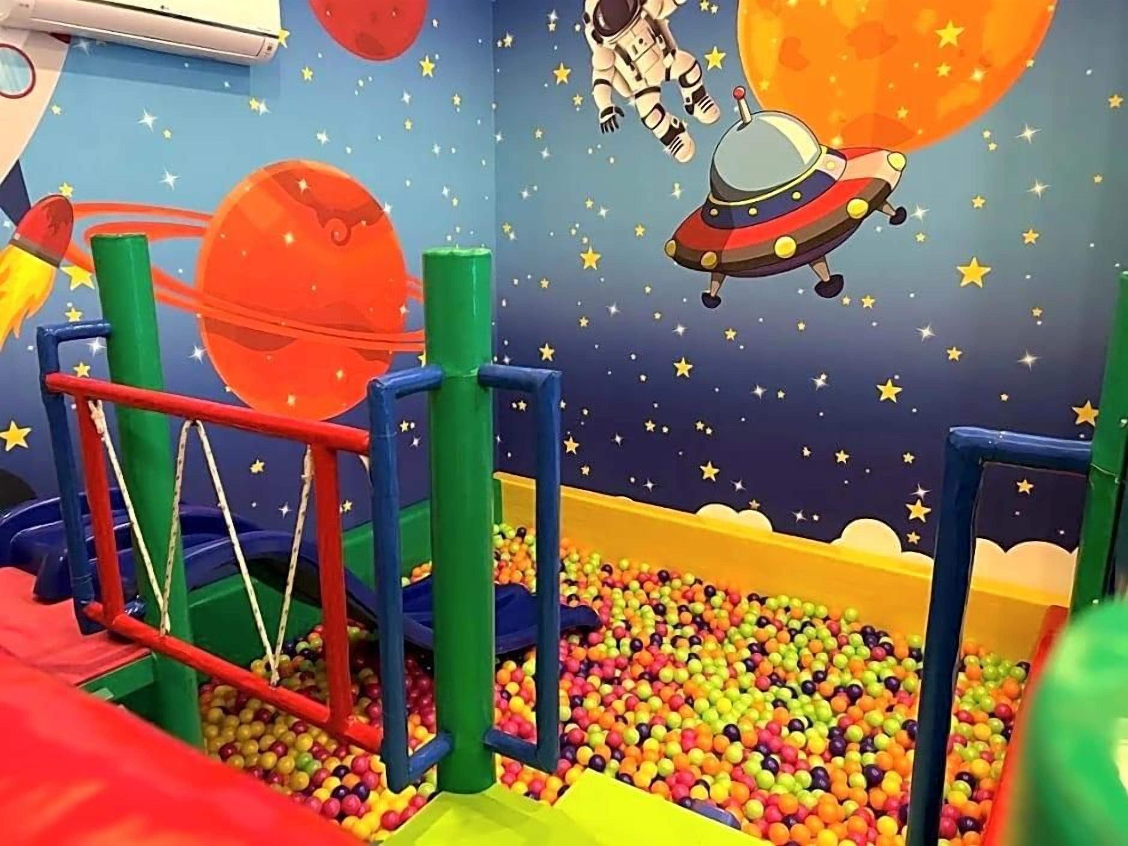 Uma área de recreação infantil com piscina de bolinhas, estruturas de jogo coloridas e um mural com tema espacial.