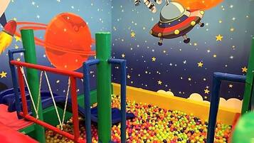 Uma área de recreação infantil com piscina de bolinhas, estruturas de jogo coloridas e um mural com tema espacial.