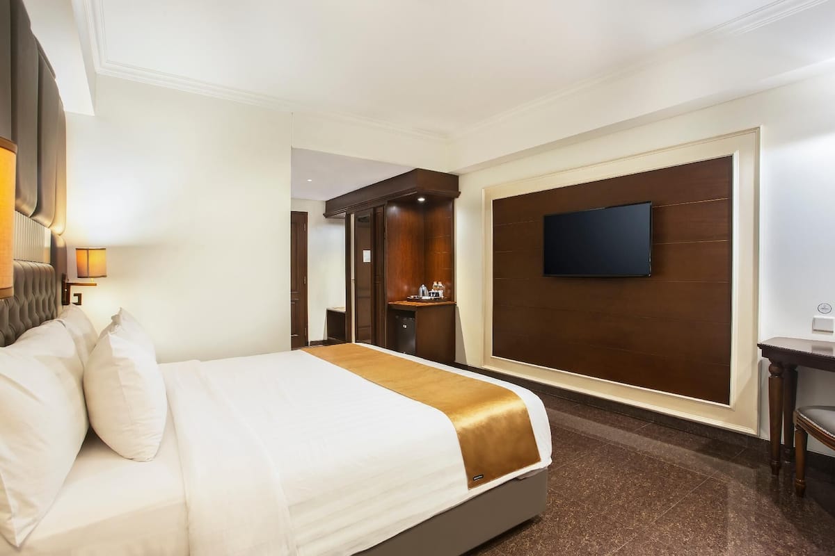 Quarto de hotel com uma cama grande, televisão de tela plana fixada na parede, uma pequena escrivaninha com cadeira e um espelho.