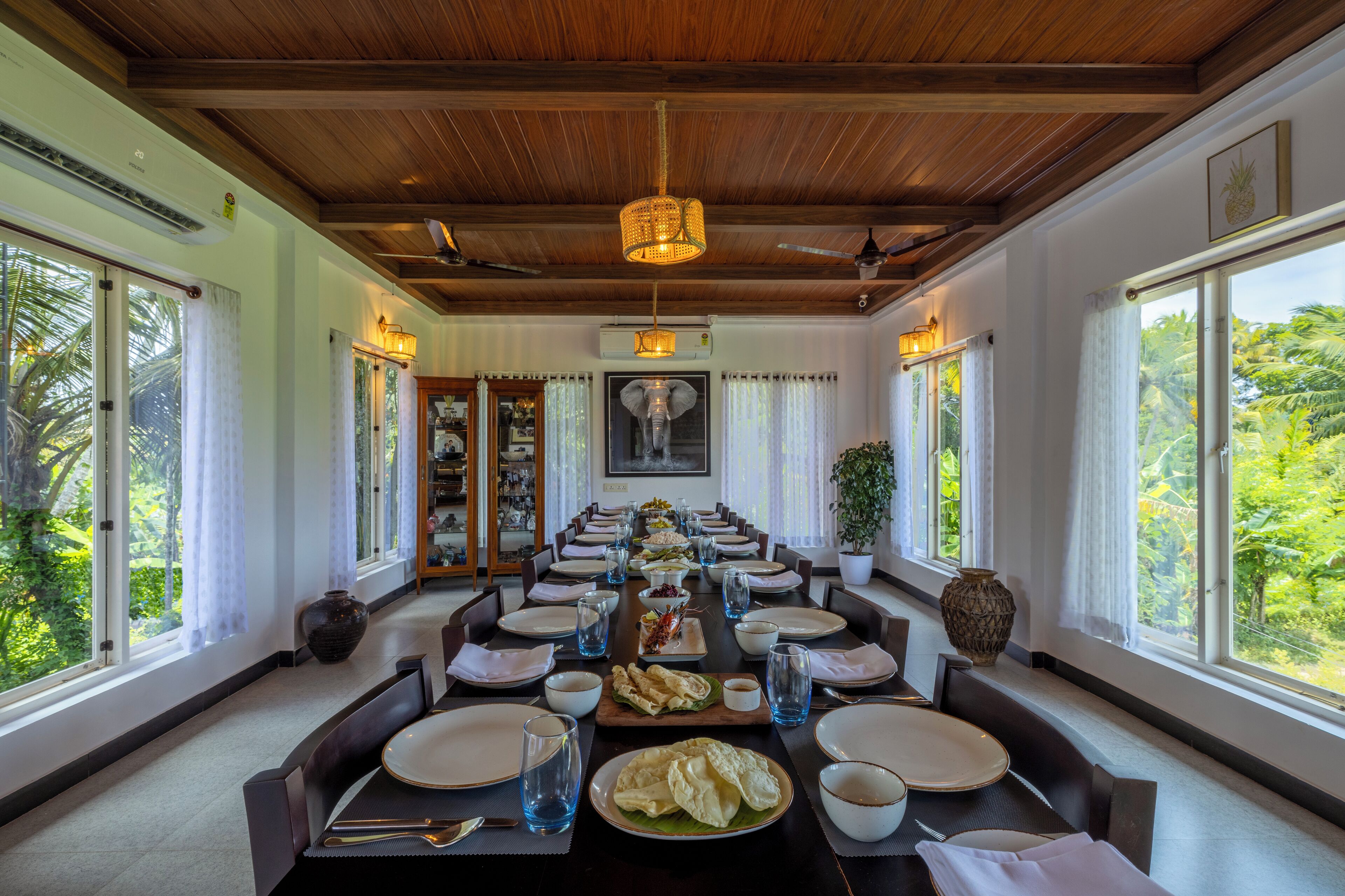 A dining area with a long table set for a meal, large windows, and a ceiling fan.