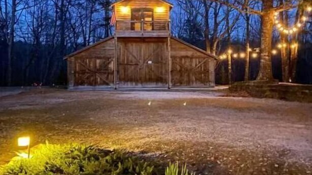 A rustic wooden cabin with a balcony and a covered porch, surrounded by trees and illuminated by string lights. - Barnaminium Upstairs Loft (Pickens)