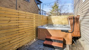 A wooden deck with a hot tub, a wooden fence, and a brick building in the background. - The Alley House - 4BR with Hot Tub (Columbus)