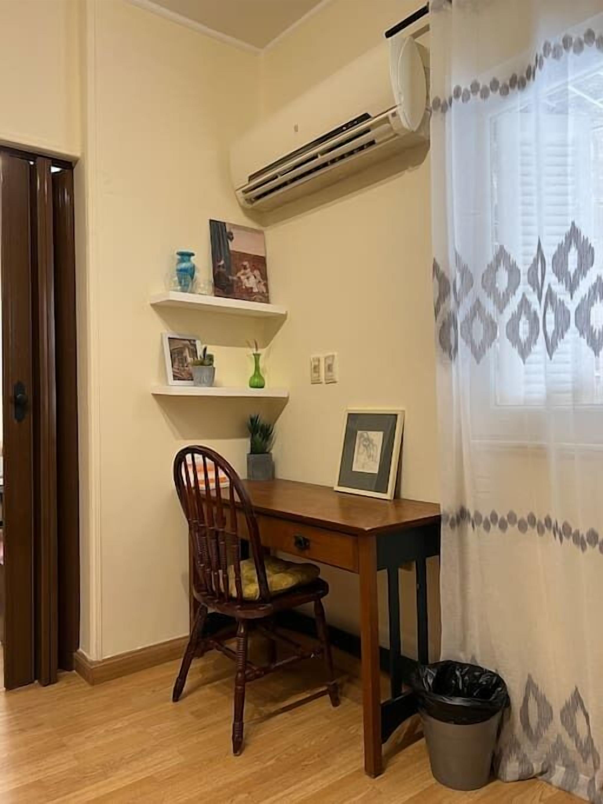A room with a wooden desk, a chair, a small trash bin, and a wall-mounted air conditioner.