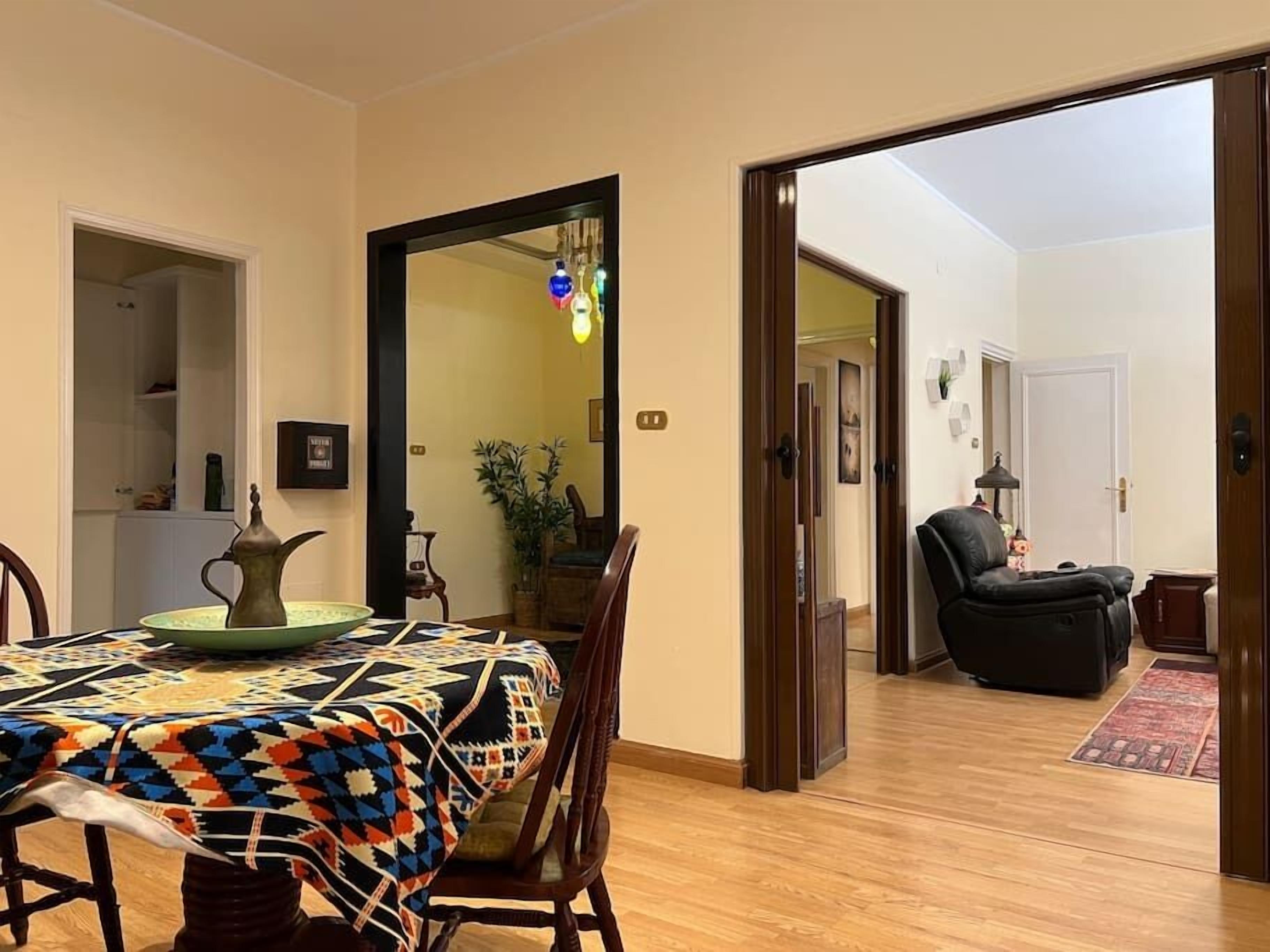 A dining area with a table set for four, a chair, and a decorative pitcher. A doorway leads to a living room with a sofa and a lamp.