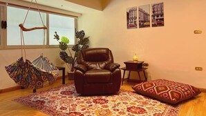 A living room with a brown leather chair, a hanging chair, a patterned rug, a potted plant, and a small table with a lamp. - Vintage Vibrant Space ! (Cairo)