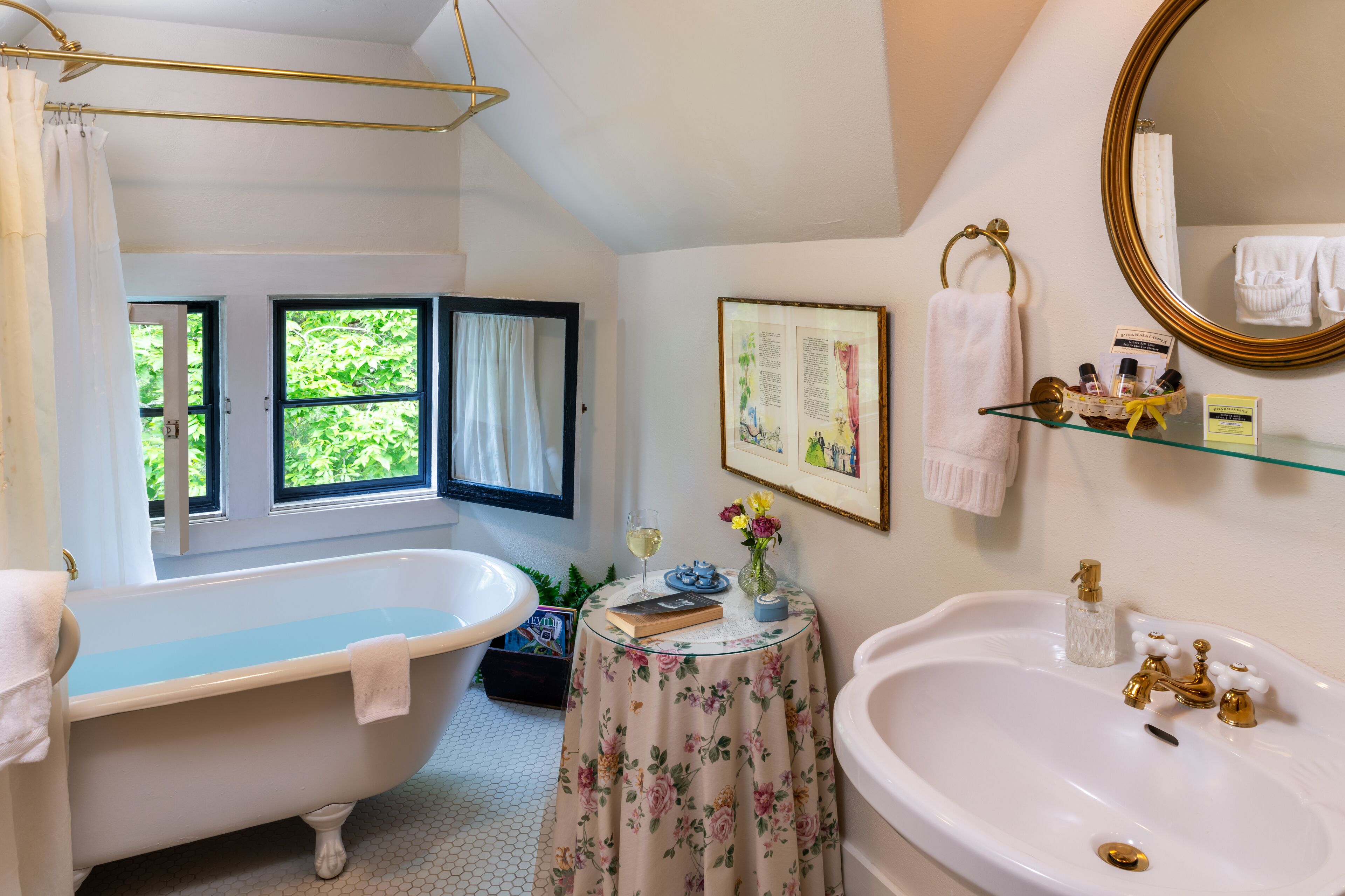 Un baño con bañera, lavamanos, ventana con cortinas, un cuadro enmarcado y una mesita con artículos de aseo.