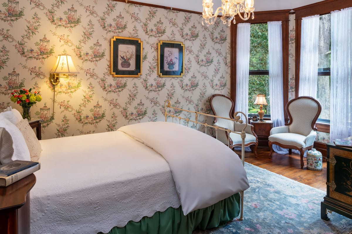 Une chambre avec un papier peint à motifs floraux, un grand lit, deux lampes fixées au mur, une chaise en bois et une vue sur la verdure par la fenêtre.