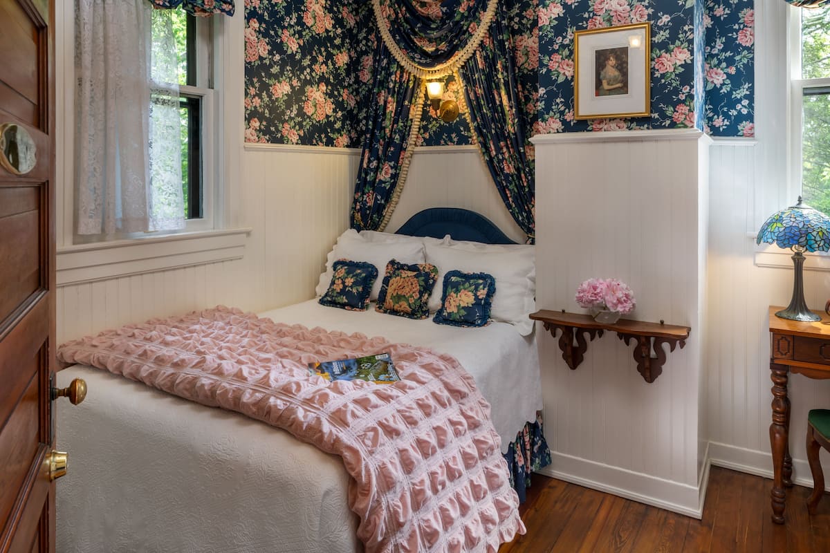 Une chambre à coucher avec un lit, un papier peint à motifs floraux, une table de chevet avec une lampe et un tableau accroché au mur.