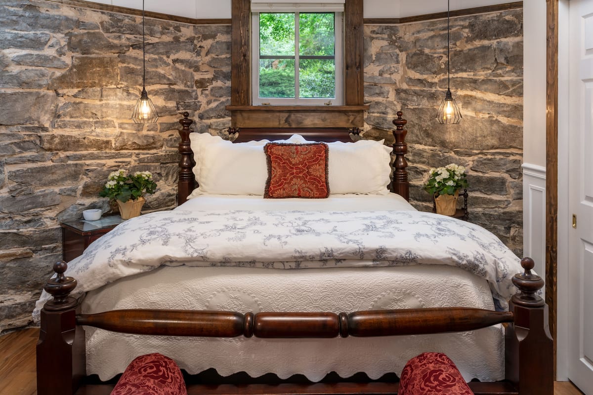 Une chambre avec un mur en pierre, un lit en bois avec une tête de lit, deux tables de chevet avec des lampes et une fenêtre donnant sur un paysage verdoyant.