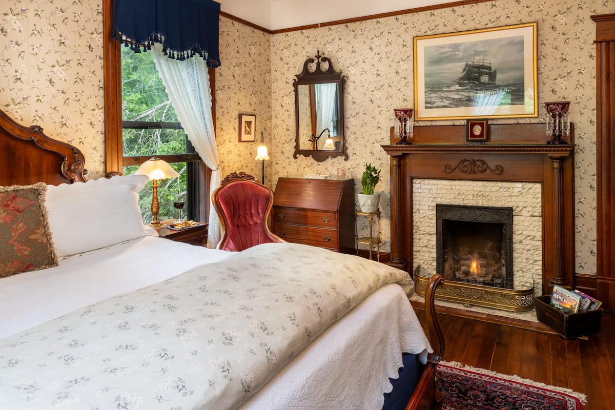 Une chambre à coucher de style traditionnel, avec un grand lit, une cheminée, un miroir et une fenêtre avec des rideaux.