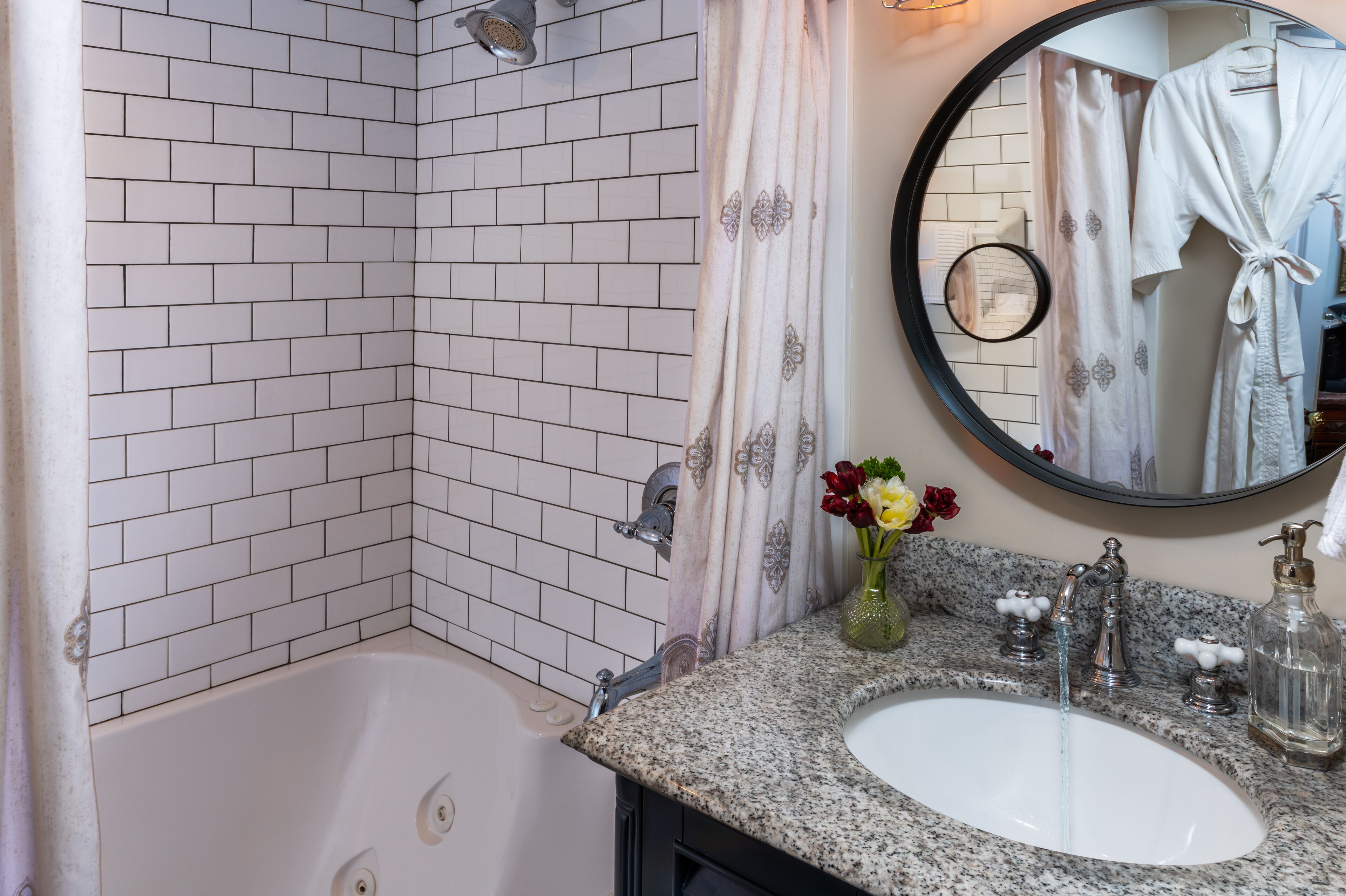A bathroom with a white tiled shower, a sink with a marble countertop, and a round mirror.