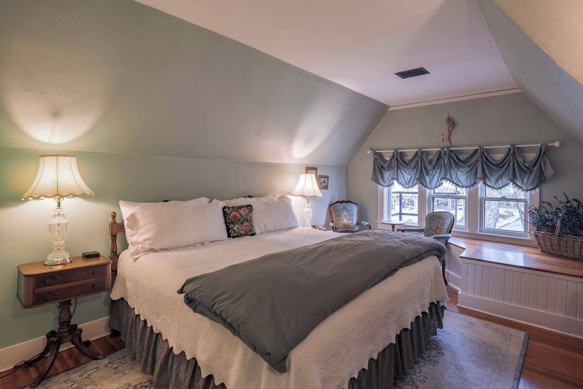 Une chambre à coucher avec un grand lit, deux lampes de chevet, une table de chevet, une chaise, une fenêtre avec des rideaux et un radiateur.