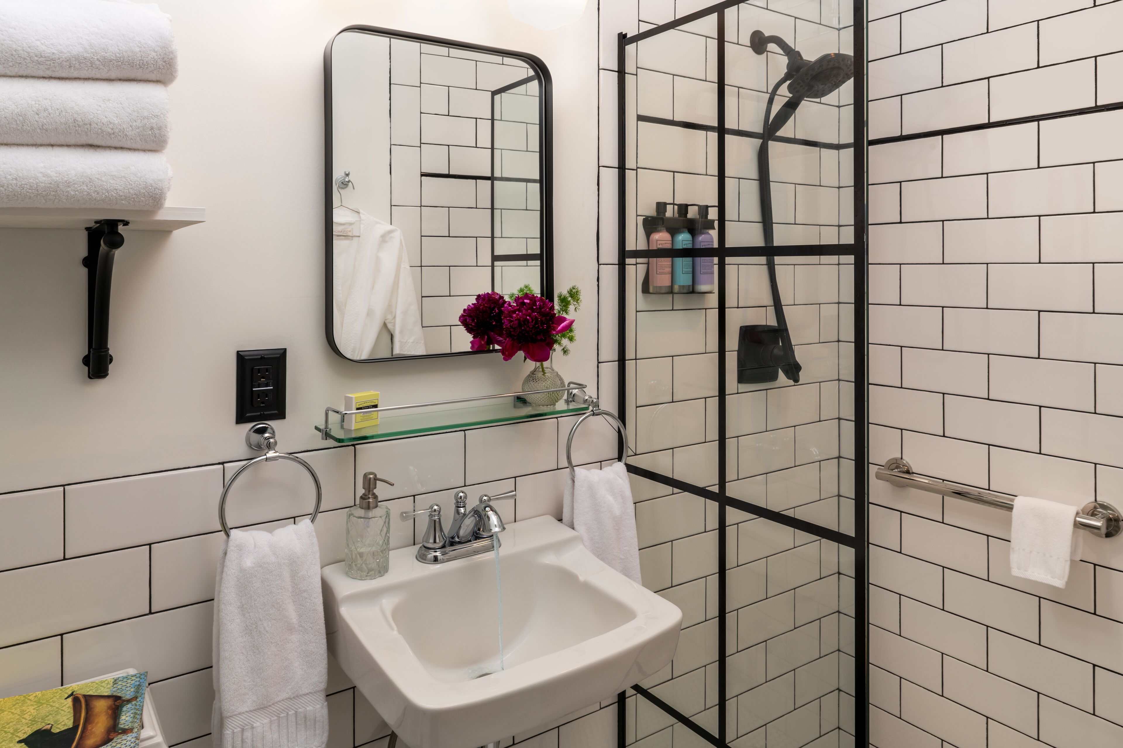Une salle de bain avec un lavabo blanc, une étagère en verre où sont rangés les articles de toilette, un miroir et un espace douche.