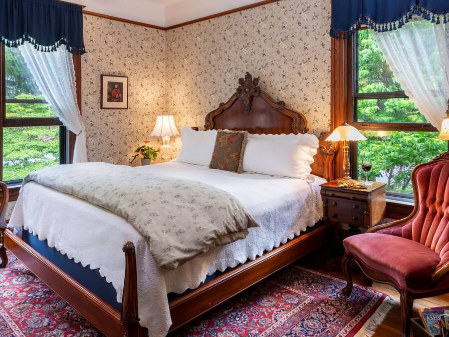 A traditional bedroom with a four-poster bed, a red upholstered chair, and a view of greenery through the windows.