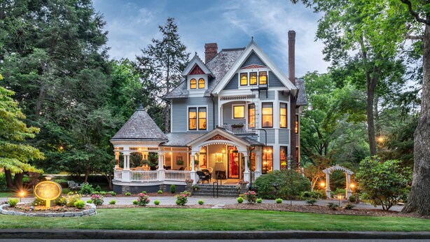 Une grande maison à deux étages, dotée d’une véranda qui fait le tour de la maison, de nombreuses fenêtres et d’une cheminée.