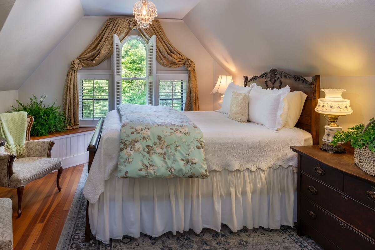 Une chambre à coucher avec un grand lit, une fenêtre avec des rideaux, un lustre, une lampe et une commode.