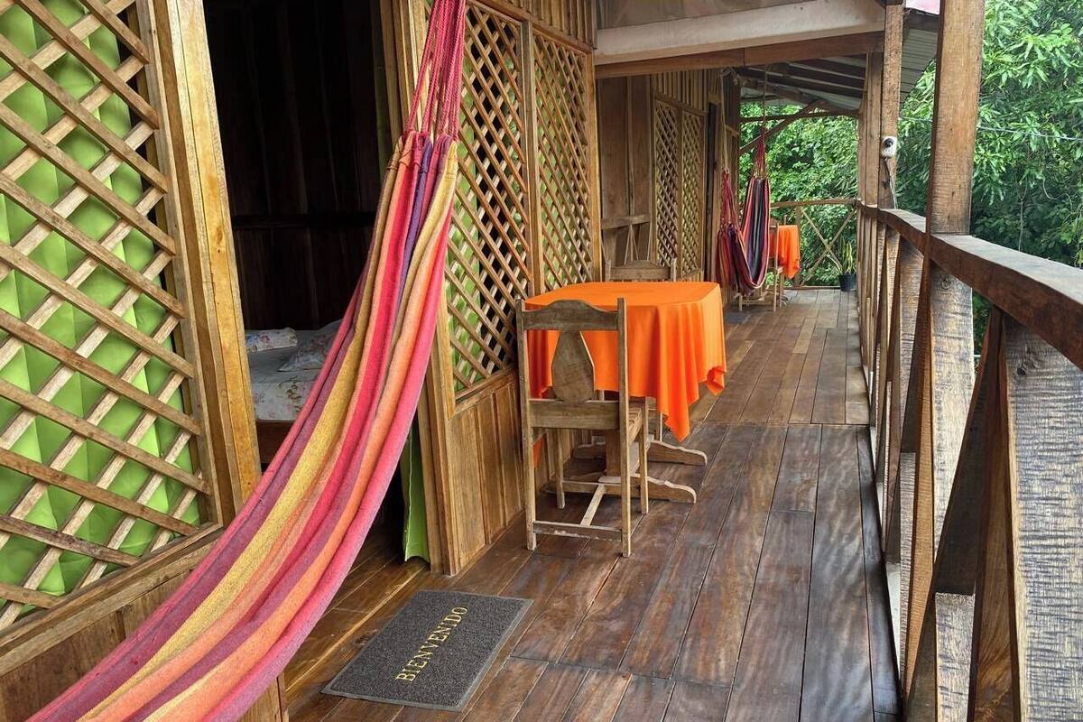 Una terraza de madera con una hamaca, una mesa con mantel naranja y una silla de madera.