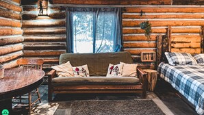 A log cabin interior with a sofa, a round table, a bed, and a window with curtains.