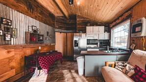 Interior rústico de cabana com teto de madeira, área de jantar com toalha xadrez, cozinha compacta com eletrodomésticos modernos e um espaço de estar aconchegante com almofada decorativa.