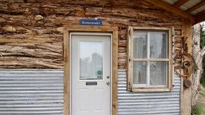 Entrada de uma cabana rústica, com porta de madeira e janela, identificada como "Homesteader - Entrada da Cabana".