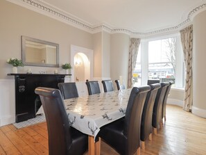Une salle à manger avec une longue table, des chaises noires, une cheminée et une grande fenêtre avec des rideaux.