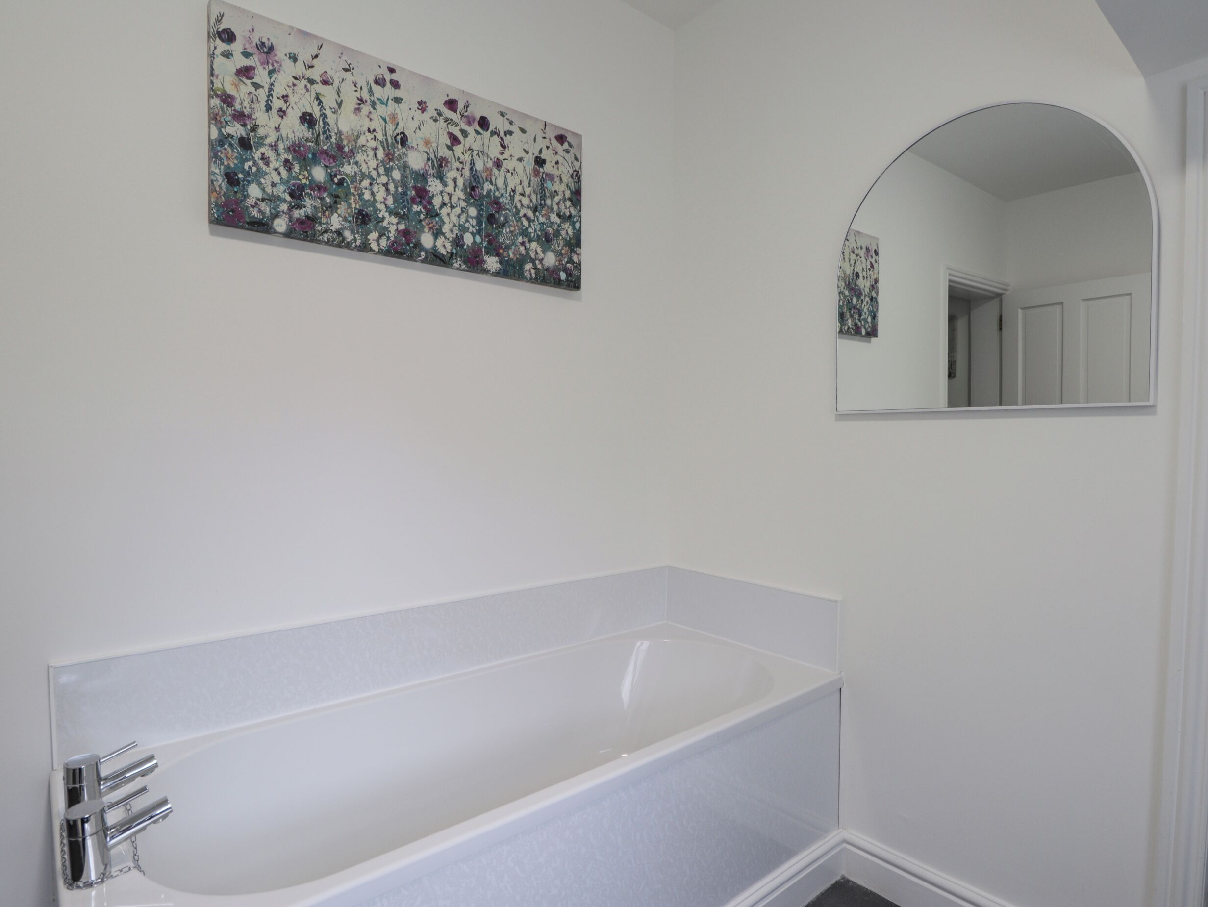 Une salle de bain avec une baignoire blanche, un miroir fixé au mur et un tableau représentant des fleurs.
