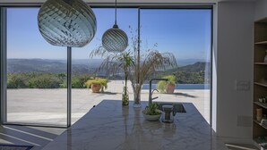 A modern kitchen with a large window, marble countertop, hanging pendant lights, and potted plants.