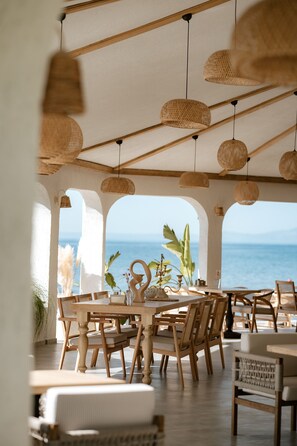 Un comedor interior con mobiliario de madera y lámparas colgantes tejidas, con vistas a un paisaje marino.