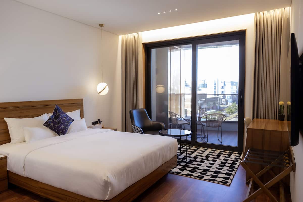 A hotel room with a large bed, a desk with a chair, a balcony with a table and chairs, and a view of the city.