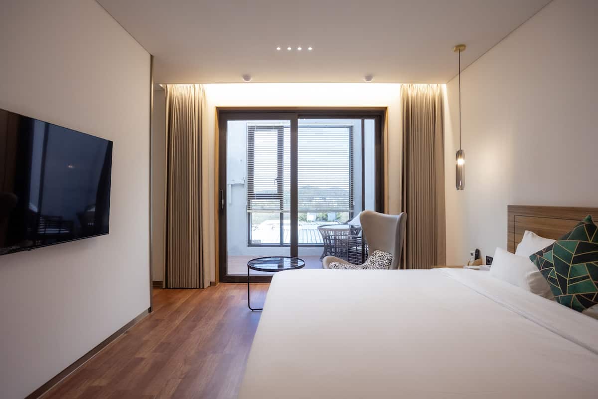A modern hotel room with a large bed, a TV mounted on the wall, a small round table, and a balcony with curtains.