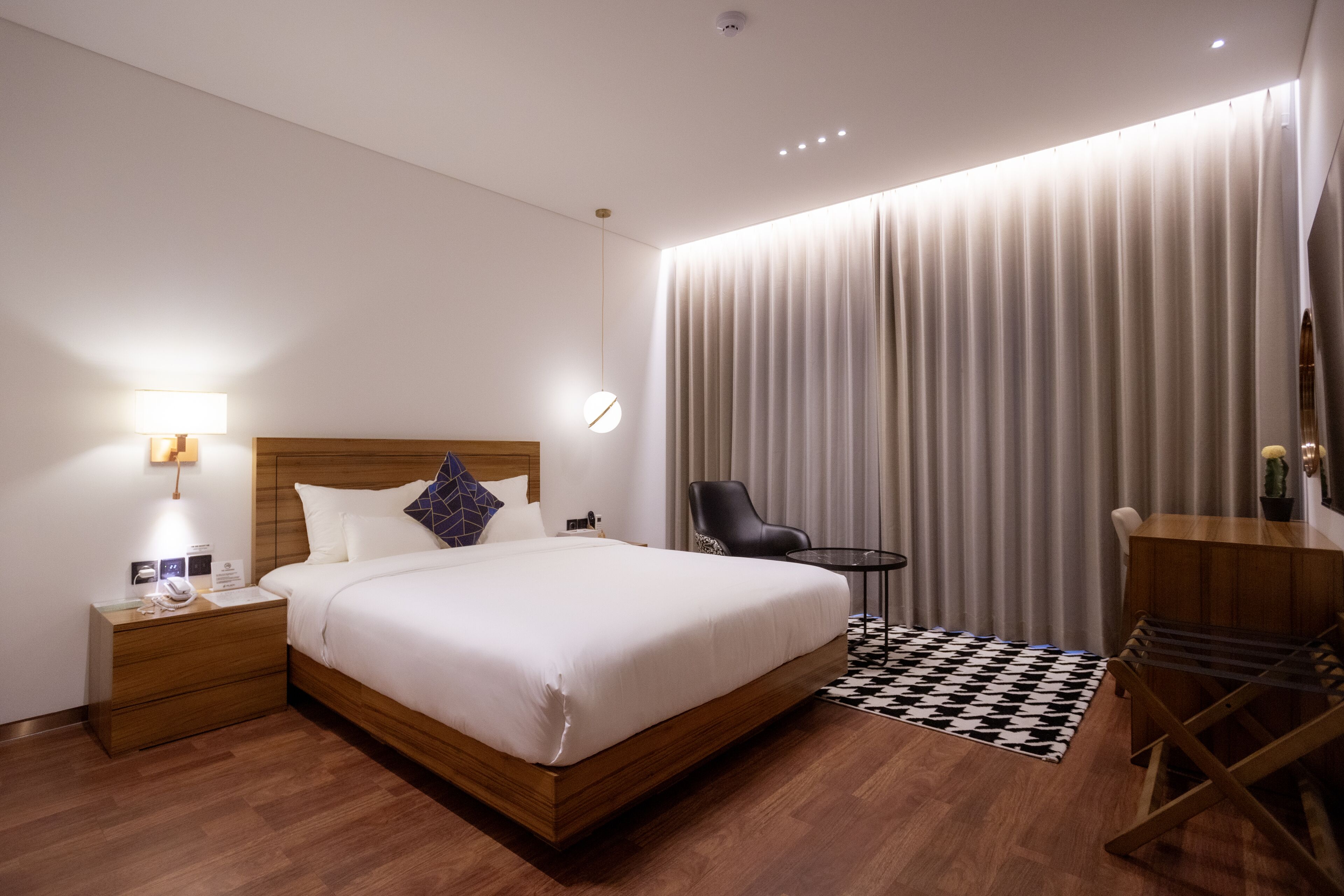A modern hotel room with a wooden bed, a small bedside table with a lamp, a chair, a desk, and a large window with curtains.