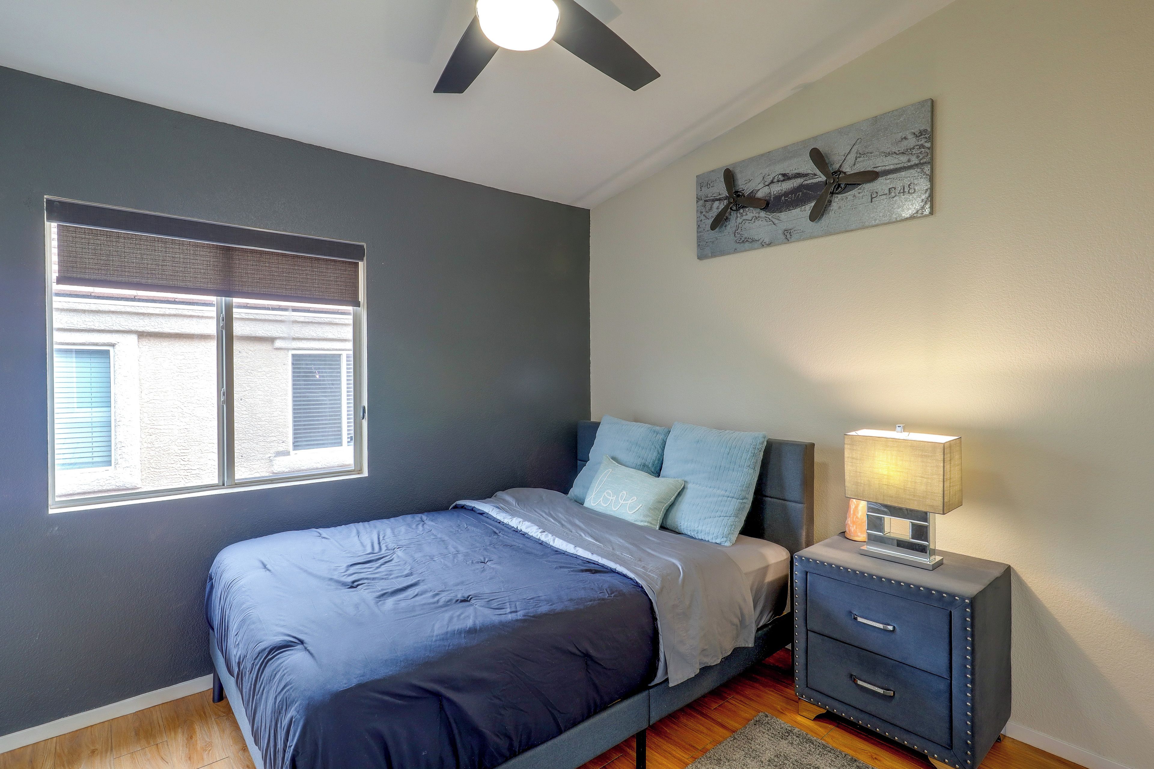 Un dormitorio con cama, mesita de noche, lámpara, ventilador de techo, ventana con persianas y una obra de arte colgada en la pared.