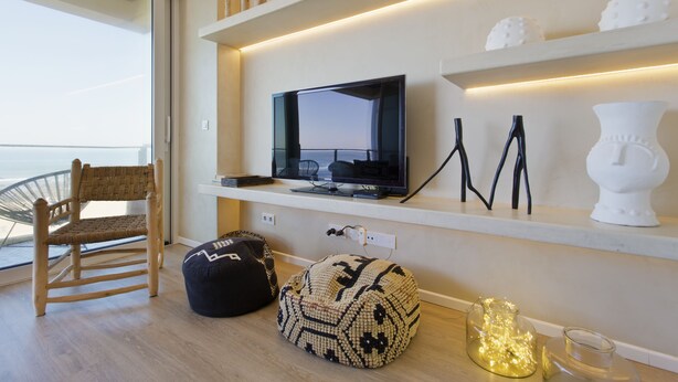 Een moderne woonkamer met een televisie op een plank, een rieten stoel, een gestreept voetenbankje en decoratieve objecten.