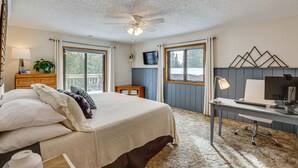 Une chambre à coucher avec un lit, un bureau avec un ordinateur, un ventilateur de plafond et de grandes fenêtres.