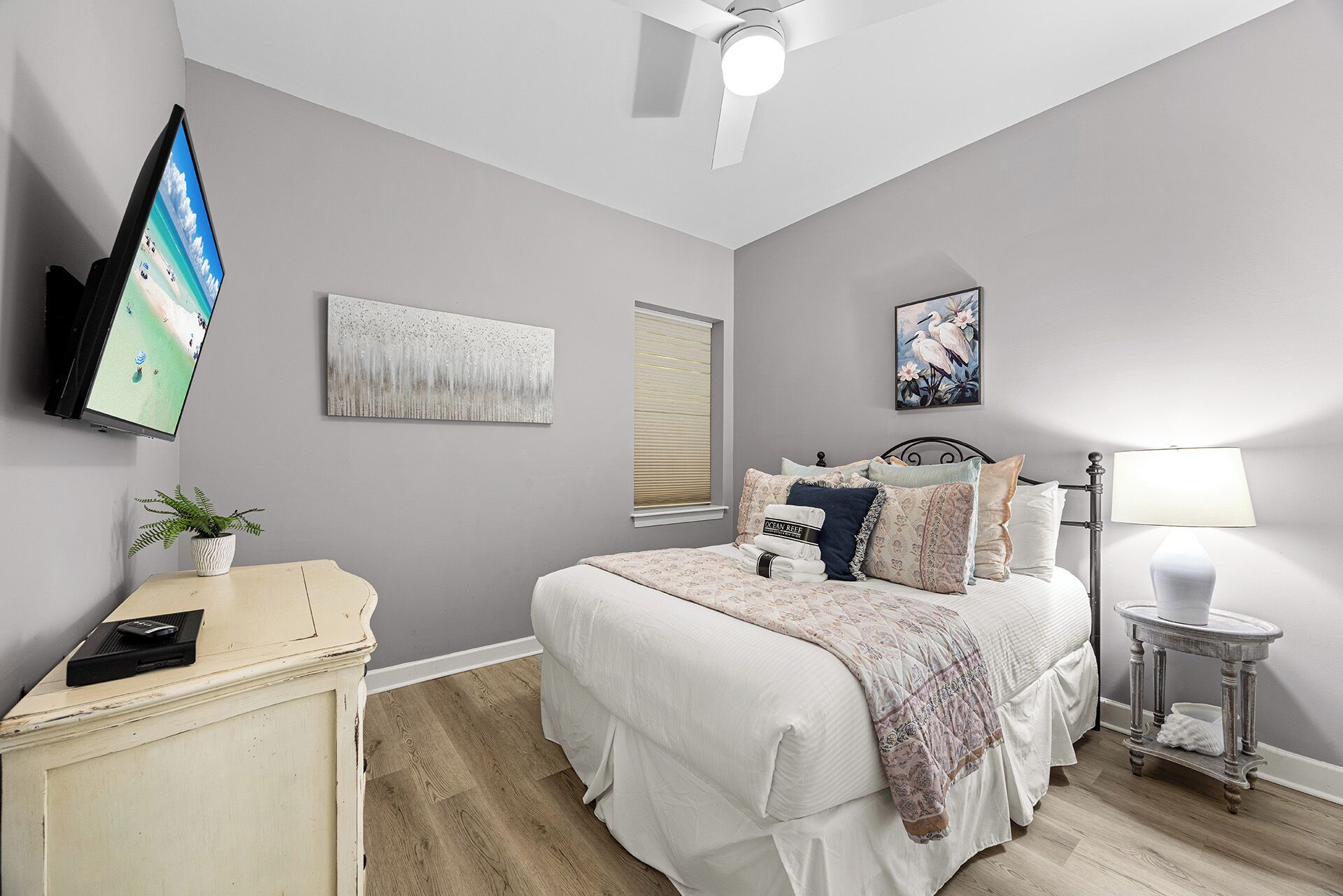 Un dormitorio con cama, televisor montado en la pared, ventilador de techo y ventana con persianas.