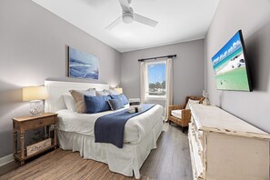 A bedroom with a bed, bedside tables, a ceiling fan, a window with curtains, and a wall-mounted TV.