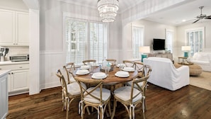 A dining area with a round wooden table set for six, surrounded by wicker chairs with cushions. A chandelier hangs from the ceiling, and a television is mounted on the wall.