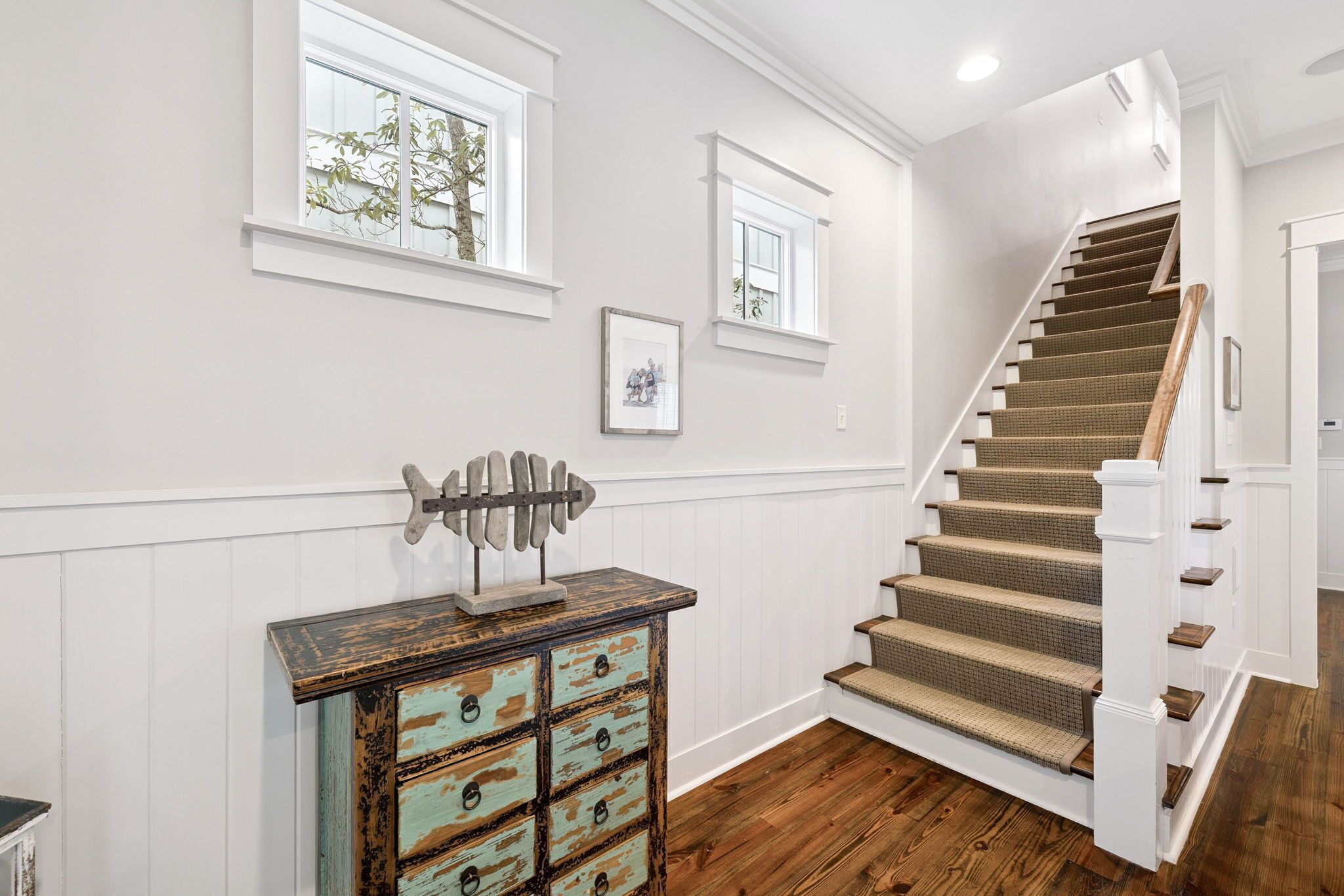 A staircase with wooden steps and a white handrail, leading to a landing with a rustic chest of drawers and framed pictures on the wall.
