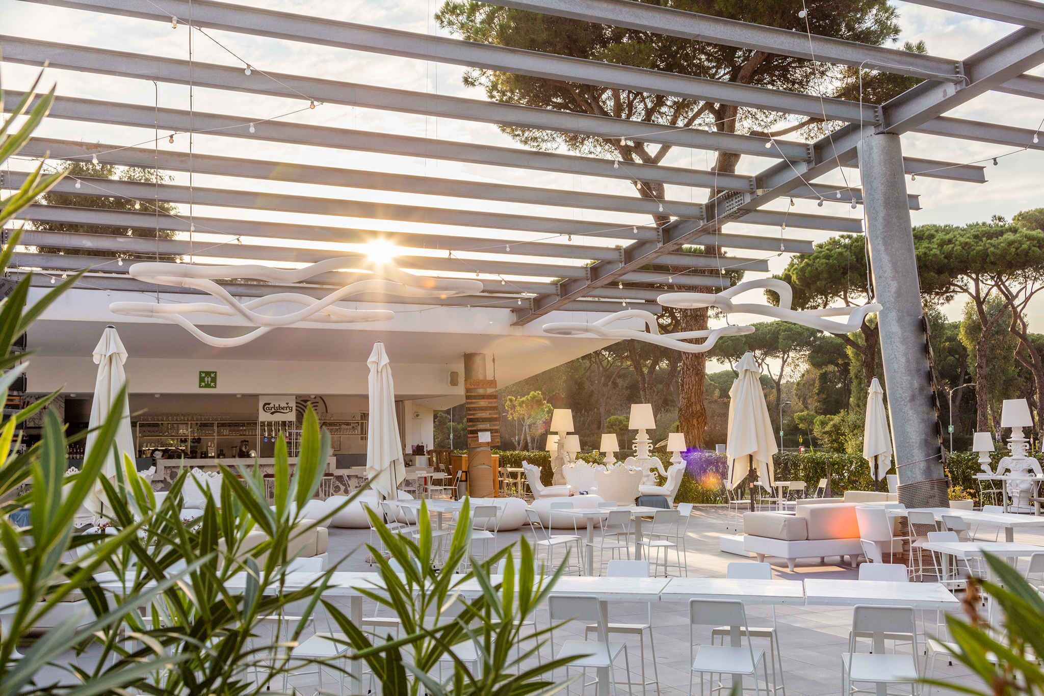 An outdoor seating area with white tables and chairs, umbrellas, and a pergola structure.