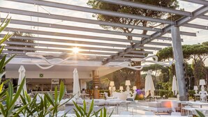 Et udendørs siddeområde med hvide borde og stole, parasoller og en pergola.